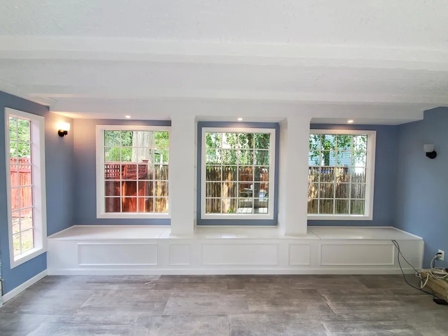 Long white built-in bench under three windows in a blue sunroom with gray plank tile flooring